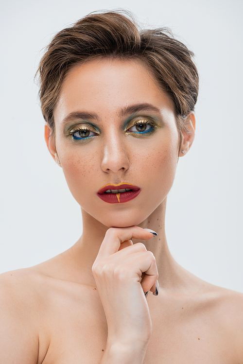 portrait of young woman with bright makeup touching chin and looking at camera isolated on grey