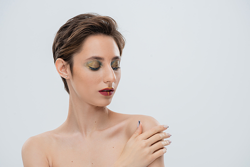 portrait of young woman with bright makeup and short hair touching bare shoulder and looking at camera isolated on grey