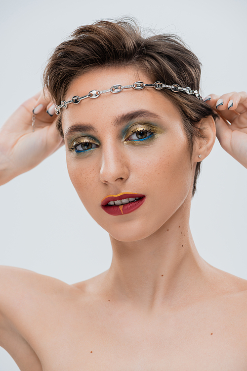 young woman with shiny makeup and short hair wearing silver chain on head isolated on grey