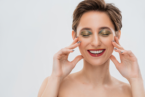 satisfied young woman with shimmery eye makeup touching cheeks isolated on grey