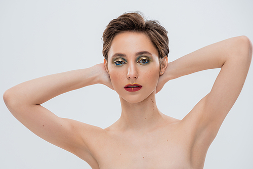 young woman with shimmery eye makeup standing with hands behind neck isolated on grey