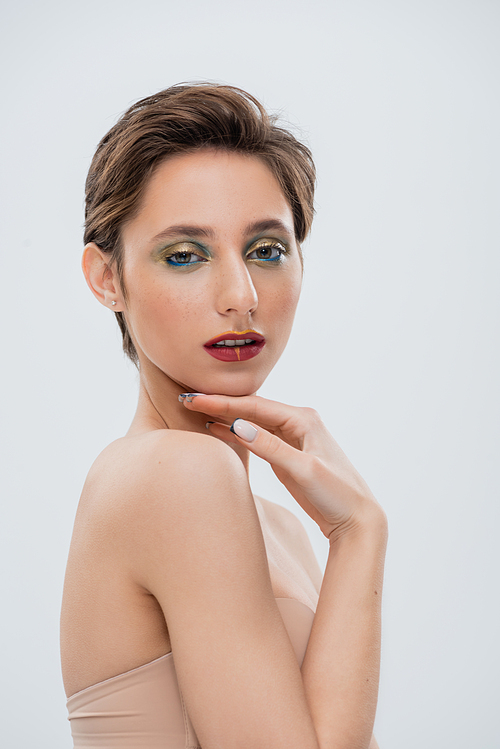 young woman with shimmery eye makeup touching chin isolated on grey