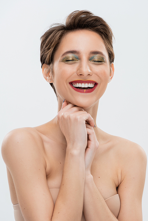 cheerful young woman with shimmery eye makeup standing in strapless bra isolated on grey