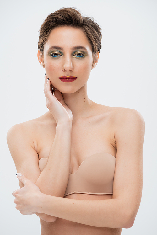 pretty young woman with shiny eye makeup standing in strapless bra isolated on grey