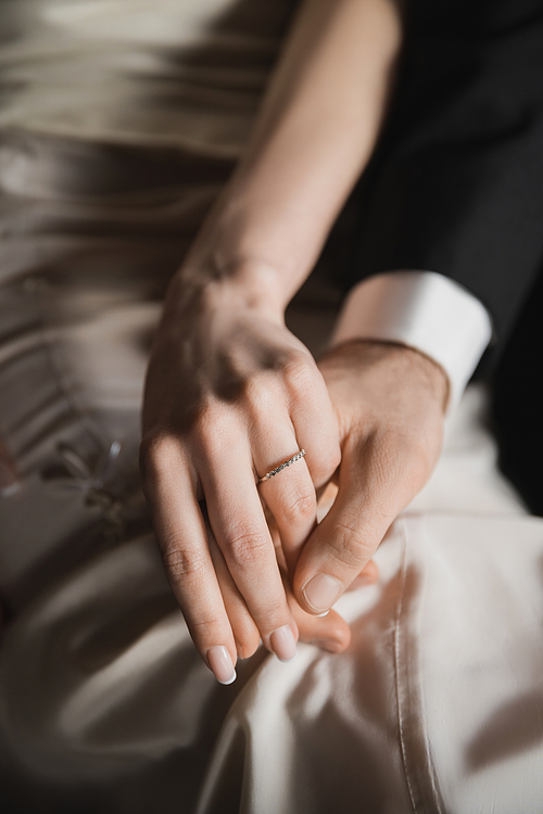 cropped view of newlyweds, bride with elegant and luxurious wedding ring on finger and groom in suit holding hands of each other after wedding in hotel room