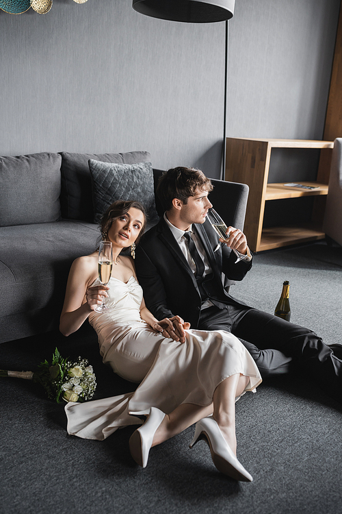 dreamy bride in elegant white wedding dress and groom in black suit holding glasses of champagne while celebrating their marriage near bridal bouquet and couch after wedding in hotel room