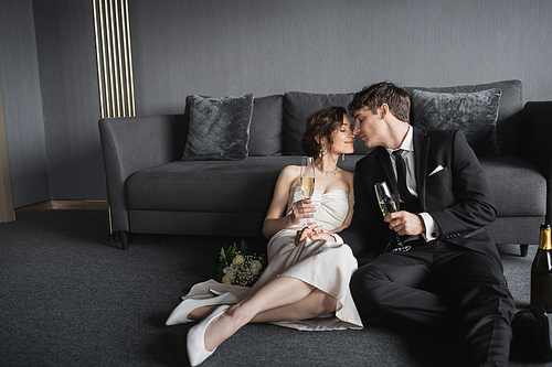 happy newlyweds, bride in white wedding dress and groom in black suit holding glasses of champagne while kissing and celebrating their marriage near bridal bouquet and couch in hotel room