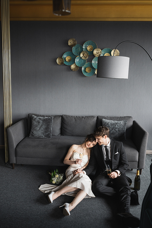 cheerful bride in wedding dress leaning on shoulder of groom in black suit and holding glasses of champagne near bridal bouquet, couch and floor lamp in hotel room