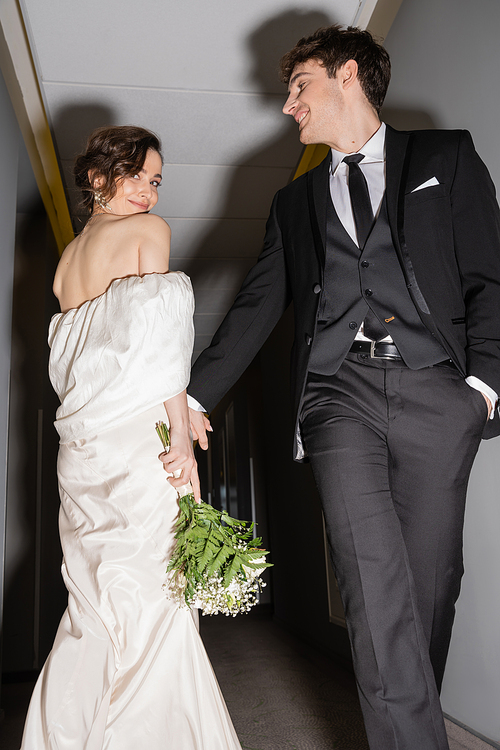 low angle view of cheerful groom in black suit posing with hand in pocket and looking at happy bride in white wedding dress holding bridal bouquet while walking together in hall of modern hotel