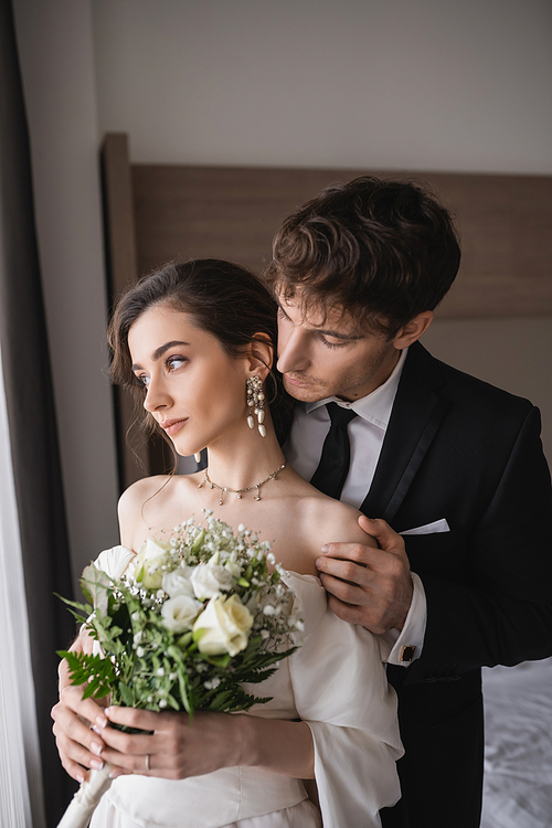 tender groom in classic formal wear touching shoulder of elegant young bride in jewelry, white dress with bridal bouquet while standing together in modern hotel room after wedding ceremony