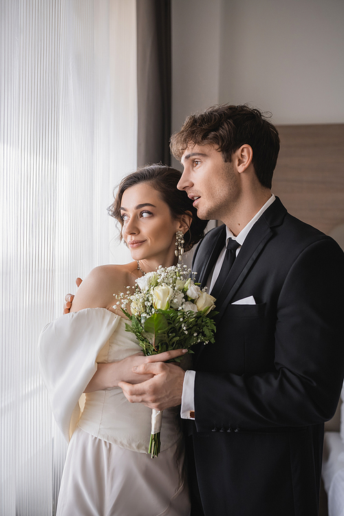 amazed groom in classic formal wear standing with opened mouth next to elegant young bride in jewelry, white dress with bridal bouquet in modern hotel room after wedding ceremony