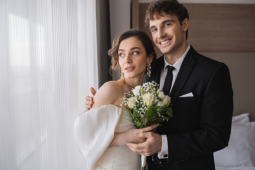 cheerful groom in classic formal wear hugging elegant bride in jewelry, white dress with bridal bouquet while standing together in modern hotel room after wedding ceremony