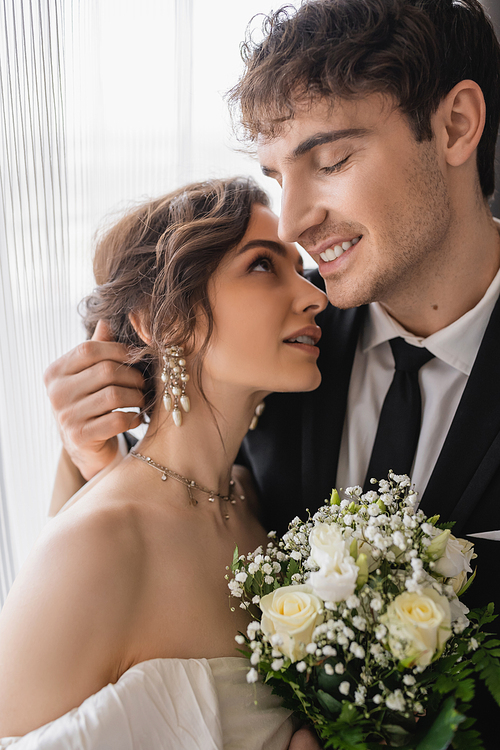 cheerful groom in classic formal wear hugging happy bride in jewelry, white dress with bridal bouquet while standing together in modern hotel room after wedding ceremony