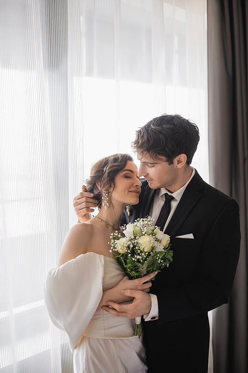 groom in classic formal wear hugging happy bride in jewelry, white dress with bridal bouquet while standing together after wedding ceremony in modern hotel room