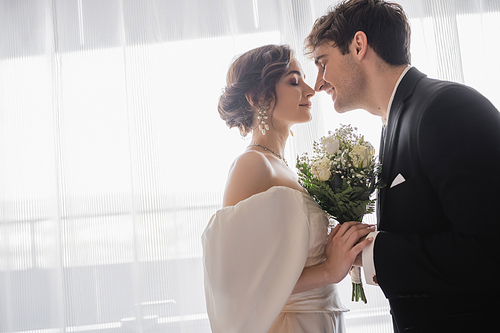 side view of young bride in jewelry, white dress with bridal bouquet and groom in classic formal wear standing together with closed eyes in modern hotel room after wedding ceremony, happy couple