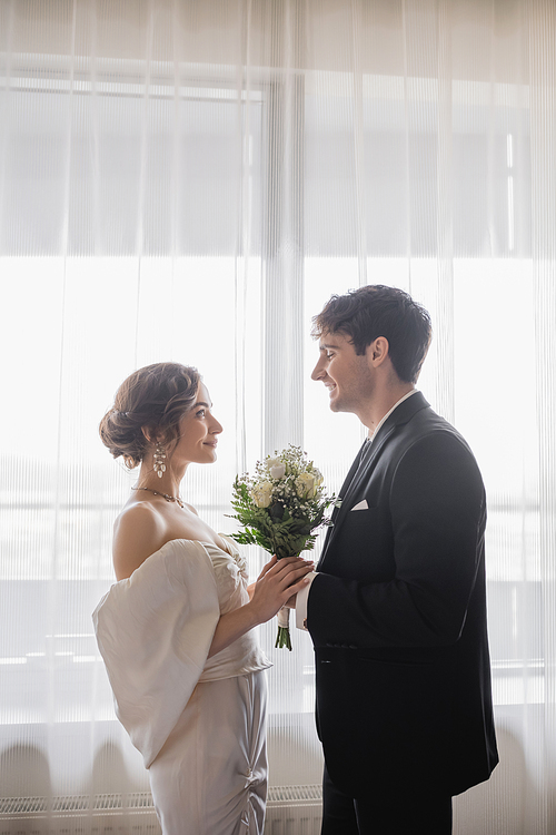 side view of bride in jewelry, white dress with bridal bouquet and groom in classic formal wear standing together and looking at each other after wedding ceremony in hotel, happy couple