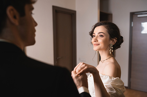 pretty young bride in elegant jewelry and white dress holding hands with blurred groom in classic formal wear while dancing together in modern hotel room after wedding ceremony, happy couple