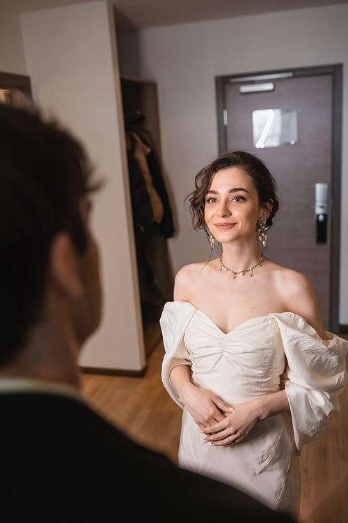 pretty young bride in elegant jewelry and white dress looking at blurred groom while standing near entrance door to modern hotel room after wedding ceremony, happy couple