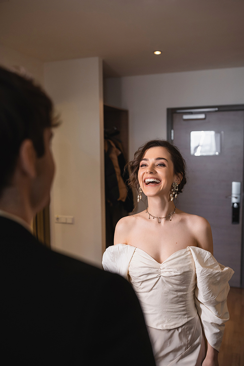 pretty young bride in elegant jewelry and white dress laughing and looking at blurred groom while standing near entrance door to modern hotel room after wedding ceremony, happy couple