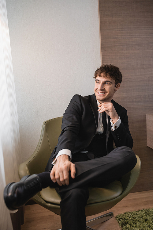 happy man in black formal wear with tie and classic shoes sitting on comfortable armchair and looking away on wedding day, resting in modern hotel room, good looking groom