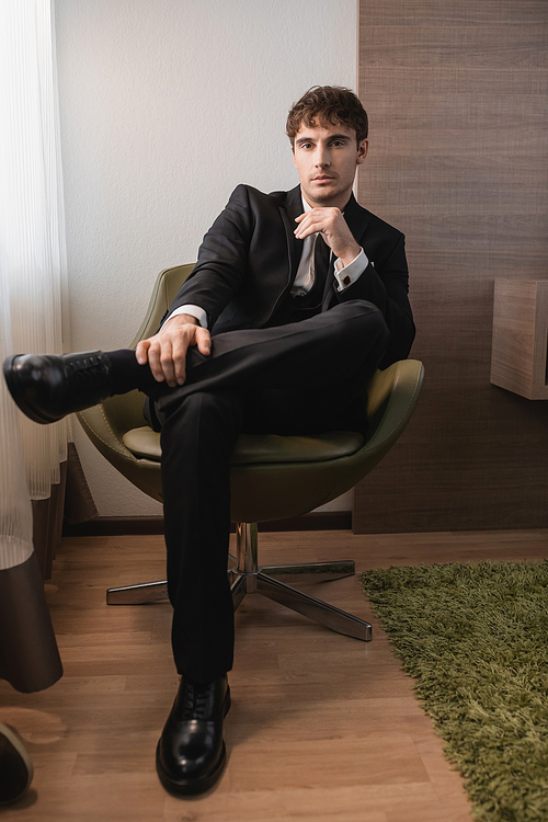 young man in black formal wear with tie and classic shoes sitting on comfortable armchair and looking at camera on wedding day, posing in modern hotel room, good looking groom