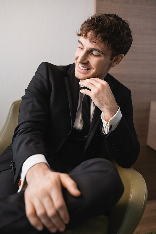 cheerful man in black formal wear with tie sitting on comfortable armchair and looking away on wedding day, resting in modern hotel room, good looking groom