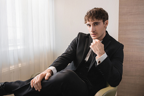 successful man in black formal wear with tie sitting on comfortable armchair and looking at camera on wedding day, resting in modern hotel room, good looking groom