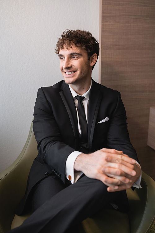 pleased man in black formal wear with tie sitting on comfortable armchair and looking away on wedding day, resting in modern hotel room before marriage, handsome groom