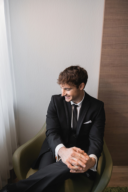 happy man in black suit with tie smiling and sitting with clenched hands on comfortable armchair and looking away on wedding day, resting in modern hotel room, good looking groom