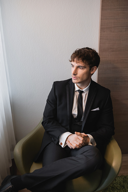 young man in black suit with tie sitting with clenched hands on comfortable armchair and looking away on wedding day, thinking in modern hotel room before marriage, good looking groom