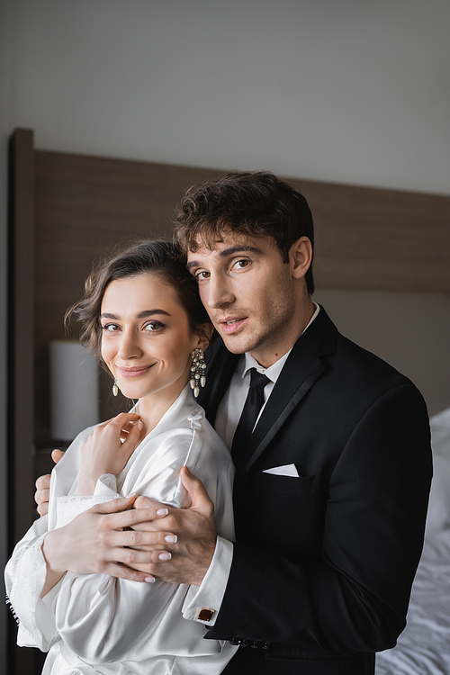 groom in classic formal wear embracing happy young bride in jewelry and white dress while standing together in modern hotel room during their honeymoon after wedding