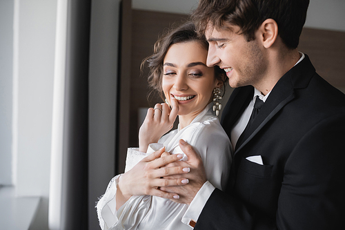 groom in classic black suit embracing happy young bride in jewelry and white dress while standing together in modern hotel room during their honeymoon on wedding day, joyful newlyweds