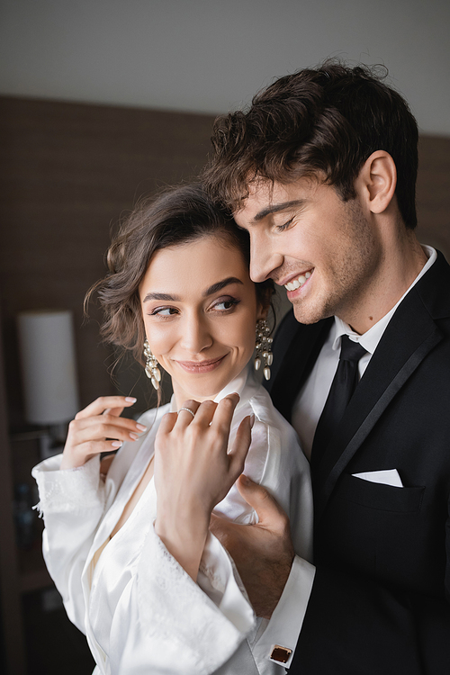 groom with closed eyed in classic black suit embracing happy young bride in jewelry and white wedding dress while standing together in modern hotel room during their honeymoon, newlyweds