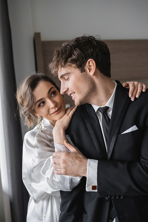 pleased bride in jewelry and white silk robe leaning on shoulder of joyful groom in classic black suit while standing together in modern hotel room during honeymoon, newlyweds