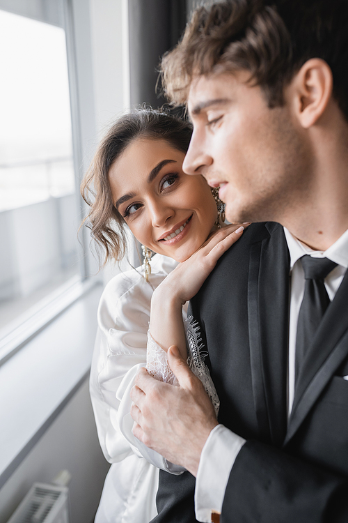 happy bride in jewelry and white silk robe leaning on shoulder of blurred groom in classic black suit while standing together in modern hotel room during honeymoon, newlyweds