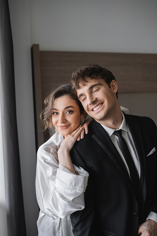 happy bride in jewelry and white silk robe leaning on shoulder of joyful groom in classic black suit while standing together in modern hotel room during honeymoon, newlyweds