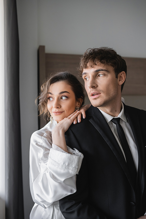 young bride in jewelry and white silk robe leaning on shoulder of groom in classic formal wear while standing and looking away together in modern hotel room during honeymoon, newlyweds