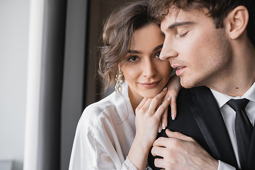 charming bride in jewelry and white silk robe leaning on shoulder of groom in classic formal wear while standing together in modern hotel room during honeymoon, newlyweds