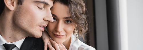 pretty bride in jewelry and white silk robe leaning on shoulder of groom in classic formal wear while standing together in modern hotel room during honeymoon, newlyweds, banner