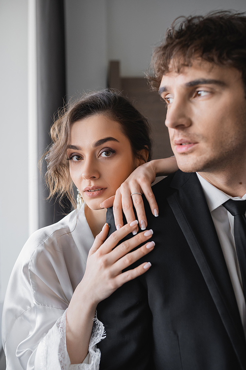 elegant bride in jewelry and white silk robe leaning on shoulder of groom in classic formal wear while standing together in modern hotel room during honeymoon, newlyweds