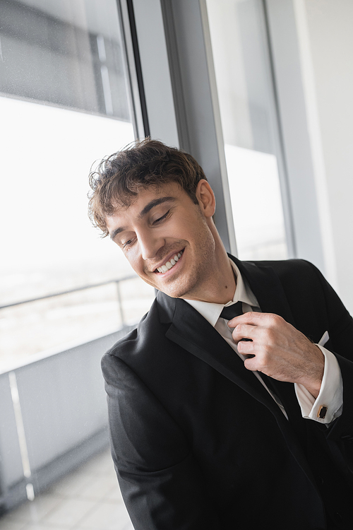 happy man in classic formal wear adjusting black tie while doing preparations and standing in modern hotel room near window, groom on wedding day, special occasion