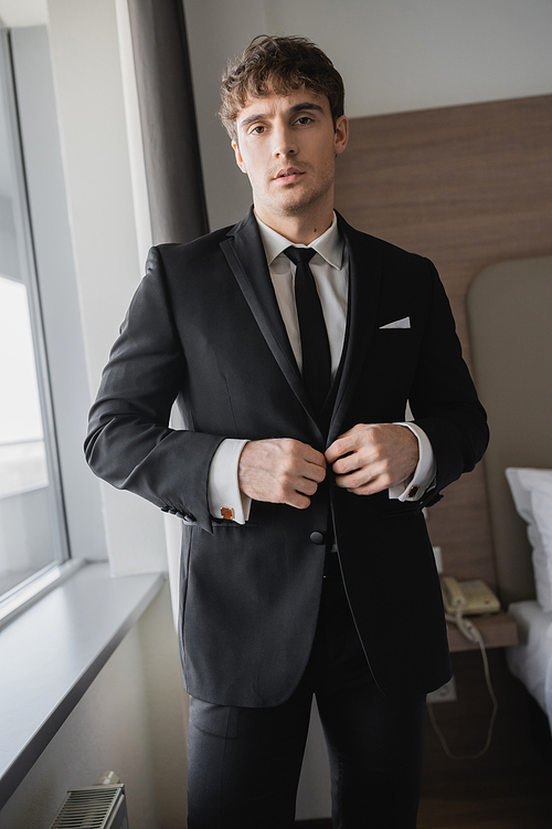 handsome man in classy formal wear with black tie and white shirt buttoning blazer and standing in modern hotel room near window, groom on wedding day, special occasion