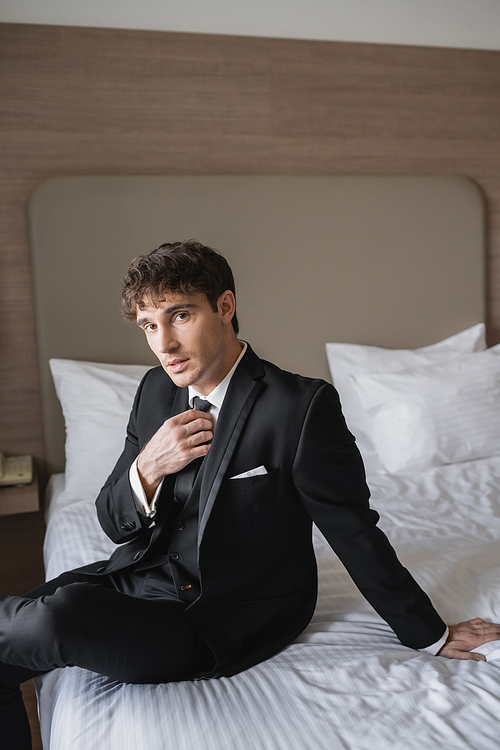 good looking man in classy formal wear with white shirt adjusting black tie while looking at camera and sitting on bed in modern hotel room, groom on wedding day, special occasion