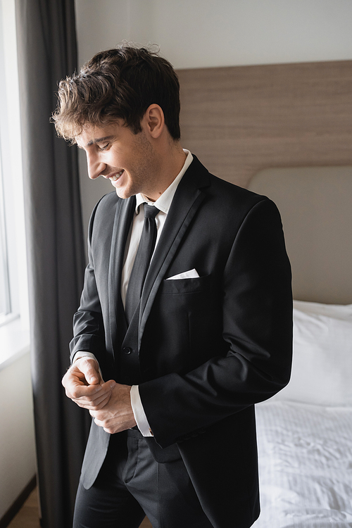 portrait of happy young man in classic black suit with tie and white shirt smiling while standing in modern hotel room, groom on wedding day, special occasion