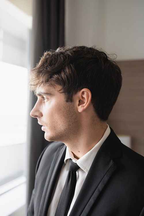 portrait of good looking man in classy formal wear with black tie and white shirt looking at window in modern hotel room, groom on wedding day, special occasion, side view