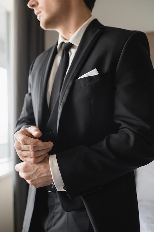 cropped view of groom in formal wear with classy black tie and white shirt standing in modern hotel room while adjusting handcuffs, groom on wedding day, special occasion