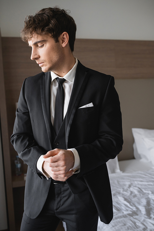 handsome groom in formal wear with classy black tie and white shirt standing in modern hotel room while adjusting handcuffs, man on wedding day, special occasion