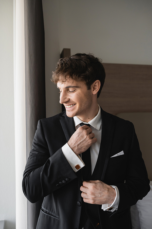 happy and good looking man in classy formal wear with white shirt adjusting black tie while smiling in modern hotel room, groom on wedding day, special occasion