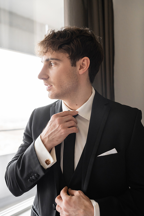 good looking groom in classy formal wear with white shirt adjusting black tie while looking at window in modern hotel room, man on wedding day, preparing for special occasion