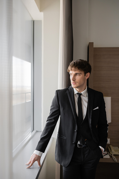 handsome man in classy formal wear with black tie and white shirt standing with hand in pocket and leaning on windowsill in modern hotel room near window, groom on wedding day, special occasion